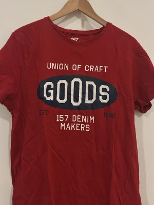 Röd t-shirt med tryck GOODS - Snygg röd t-shirt med vit och marinblå text på bröstet där det står 'Union of Craft GOODS 157 Denim Makers'. Klassisk rund hals och korta ärmar. Perfekt för en avslappnad stil.