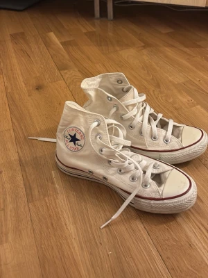 Vita Converse All Star high top sneakers - Klassiska vita Converse All Star high top sneakers med vit canvas, vita snören och gummisula. Skorna har den ikoniska blå och röda loggan på sidan och metallöljetter för snörningen. Perfekta för en avslappnad och cool streetstil.