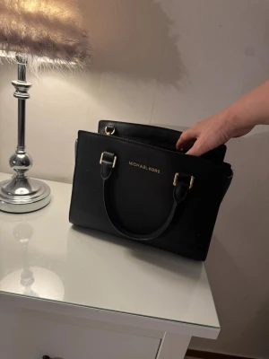 Svart handväska från Michael Kors - Snygg svart handväska från Michael Kors i strukturerat läder med guldfärgade metalldetaljer. Väskan har två handtag och en stilren, fyrkantig form. Perfekt för dig som vill ha en klassisk och trendig accessoar.