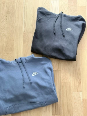 Nike hoodies i svart och blått - strl S - Två stilrena hoodies från Nike, en i svart och en i blått. Båda har klassisk huva, magficka och Nike-logga broderad på bröstet. Perfekta för en avslappnad och sportig look. Tillverkade i mjukt bomullsmaterial.