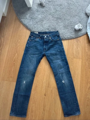 Blå Levis 501 jeans - Säljer ett par klassiska blå jeans med raka ben och snygga slitningar på både fram- och baksida. Jeansen har fem fickor, knappgylf och kontrastsömmar. Perfekta för en avslappnad och trendig look.