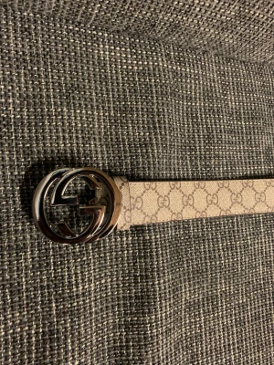 Beige bälte - Säljer ett beige bälte med klassiskt mönster och ett stort, rundat metallspänne med logga. Bältet har en stilren design och passar perfekt till jeans eller byxor för att lyfta din outfit.