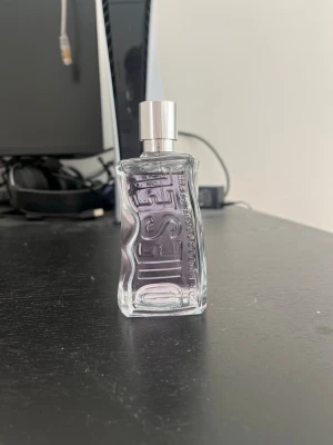 Diesel parfym i vågig glasflaska - Snygg parfym från Diesel i en transparent, vågformad glasflaska med silverfärgad spraykork. Flaskan har Diesel-loggan i relief på framsidan och en modern, stilren design som sticker ut i badrumshyllan.