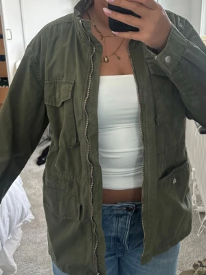 Olivgrön vindjacka med dragkedja - Snygg olivgrön vindjacka med flera fickor och dragkedja framtill. Jackan har en ståkrage och är tillverkad i ett lätt material, perfekt för lager på lager. Passar till jeans och ger en avslappnad streetstyle-look.