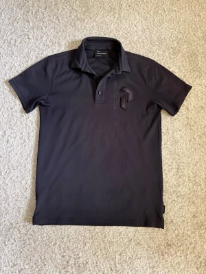 Peak Performance Golf Piké - Peak Performance golfpiké i mörkblå färg med broderad logga på bröstet. Funktionsmaterial som passar perfekt på golfbanan men fungerar lika bra till vardags. Mycket fint skick.  ✔️ Storlek S ✔️ Golfpiké ✔️ Funktionsmaterial ✔️ Mycket fint skick  📦 Skickas snabbt!