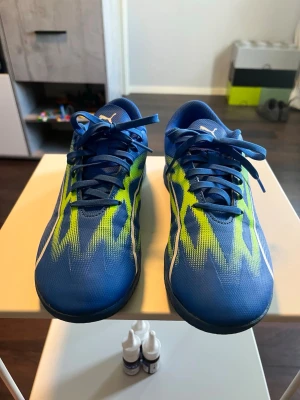 Blåa Puma fotbollsskor med neonmönster - Snygga blåa fotbollsskor från Puma med neonlimegröna och vita detaljer. Skorna har en låg profil, klassisk snörning och är tillverkade i ett lätt syntetmaterial som ger bra känsla på planen. Perfekta för dig som vill sticka ut på fotbollsplanen.