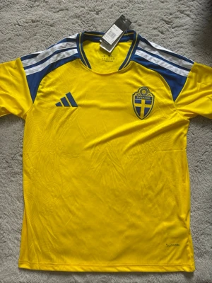 Svensk landslagströja hemma Adidas  - Säljer en officiell svensk landslagströja från Adidas i gult med blå detaljer och vita ränder på axlarna. Tröjan har korta ärmar, broderad landslagssköld och Adidas-logga på bröstet. Tillverkad i lätt och ventilerande funktionsmaterial.