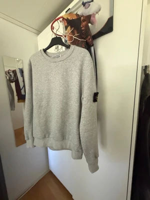 Grå sweatshirt från Stone Island - Säljer en stilren grå sweatshirt från Stone Island med den klassiska svarta patchen med gul brodyr på ärmen. Tröjan har rund halsringning, ribbade muddar och är gjord i mjukt bomullsmaterial. Perfekt för en clean och avslappnad look.