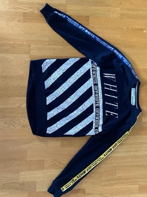 Svart sweatshirt från Off-White - Svart sweatshirt från Off-White med vita diagonala ränder och sprucken effekt på både fram- och baksida. Texten 'WHITE' och 'OFF-WHITE' syns tydligt. Ärmarna har blått och gult band med Off-White-logga. Rund hals och lång ärm.