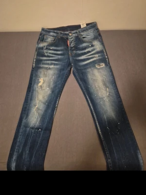 Blå slitna jeans med distressed detaljer - Säljer ett par blå jeans med coola slitningar och distressed detaljer framtill. Jeansen har en klassisk femficksmodell och raka ben. Materialet är denim och färgen är tvättad blå med ljusare partier och små färgstänk för en trendig look.