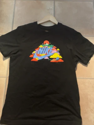 Svart Nike t-shirt med färgglatt tryck - Säljer en svart t-shirt från Nike med ett stort färgglatt tryck på bröstet som visar ett landskap med moln, träd och en glad sol. T-shirten har rund hals och korta ärmar. Perfekt för dig som gillar streetwear och vill sticka ut.