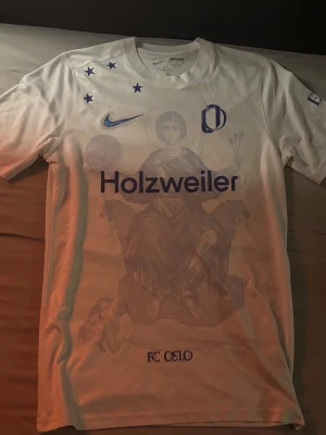 Vit fotbollströja, FC Oslo - Vit fotbollströja med blå tryck, inklusive Holzweiler-logga, Nike-logga och flera stjärnor. T-shirten har även ett unikt konstnärligt motiv på framsidan. Perfekt för dig som gillar streetwear och sportiga detaljer.