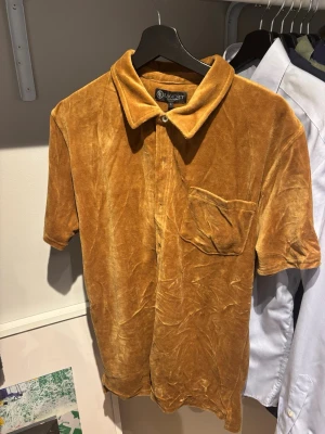 Brun frottéskjorta från Lagency - Helt ny brun skjorta i mjuk sammet/frotté från Lagency, prislapp kvar. Skjortan har korta ärmar, klassisk krage och en bröstficka. Perfekt för dig som vill sticka ut med en retrovibe och skön känsla. Passar till många olika stilar.