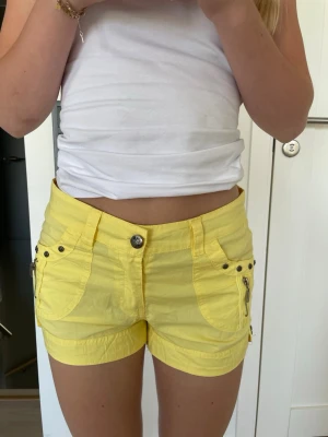 Gula shorts med coola detaljer - Snygga gula shorts med coola detaljer som dragkedjor och nitar vid fickorna. Shortsen har bälteshällor och knappstängning framtill. Perfekta för sommaren 