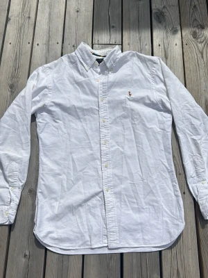 Vit skjorta med broderad logga - Klassisk vit skjorta i bomull med knappar framtill och button-down krage. På bröstet finns en liten broderad logga med en ryttare. Skjortan har lång ärm och en tidlös, normal passform. Perfekt till jeans eller chinos.