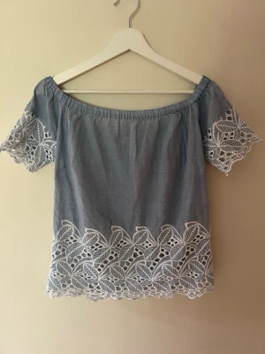 Blå offshoulder topp med spetsdetaljer - Söt blå offshoulder topp med korta ärmar och vita spetsdetaljer längs ärmslut och nederkant. Toppen har en avslappnad passform och elastisk kant upptill för att sitta snyggt på axlarna. Perfekt för varma dagar och en trendig look. Står att den är storlek M/38 men skulle säga att det mer är som en S/36. Knappt använd så den är i väldigt bra skick.