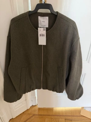 Olivgrön bomberjacka från Mango - Snygg olivgrön bomberjacka från Mango med rund halsringning och dragkedja framtill. Jackan har två snedställda fickor och elastisk nederkant för en avslappnad look. Perfekt för dig som gillar stilrena och enkla plagg. Helt ny med prislapp kvar 