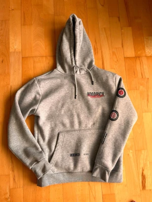 Ljusgrå hoodie från Hoodrich - Snygg ljusgrå hoodie från Hoodrich med stor huva och dragsko. Tryck på bröstet, ärmen och ryggen i svart och rött. Klassisk känguruficka framtill och ribbade muddar. Perfekt för en chill och street-inspirerad look.