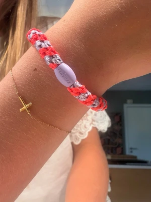 Färgglatt hårband från Kknekki - Säljer ett snyggt armband från Kknekki i flätad design med neonrosa och silverfärgade trådar. Armbandet har en lila pärla med Kknekki-loggan ingraverad. Perfekt accessoar för att addera färg till din stil.