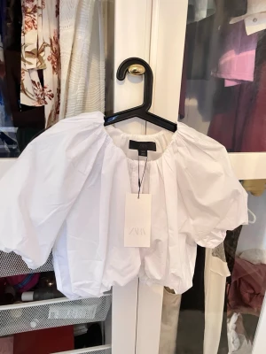 Vit puffärmad croppad topp från Zara - Nytt med prislapp! Supersnygg vit croppad topp från Zara med puffiga korta ärmar och resår i nederkant och halsringning. Perfekt för dig som vill ha en trendig och luftig look. Tillverkad i bomull för en skön känsla. 