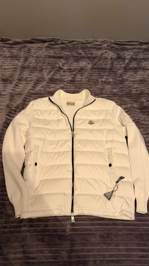 Vit dunväst från Moncler - Snygg vit dunväst från Moncler med stickade ärmar och ribbad krage. Västen har dragkedja framtill, två sidofickor och Moncler-logga på bröstet. Perfekt för lager-på-lager och ger en clean look till din outfit.