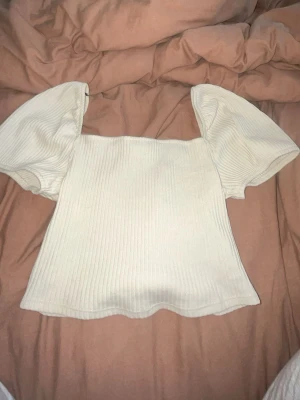 Vit ribbad topp med puffärm - Supersöt vit ribbad topp med puffärmar och fyrkantig ringning. Toppen har en croppad passform och är perfekt för dig som gillar en trendig och stilren look. Tillverkad i mjukt material som känns skönt mot huden.