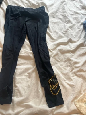 Svarta leggings från Nike med guldtryck - Snygga svarta leggings från Nike med ett stort guldfärgat Nike-tryck längs ena benet. De har en klassisk tight passform och resår i midjan. Perfekta för dig som vill ha en sportig och trendig look. Dom är för korta så det är därför jag säljer. 