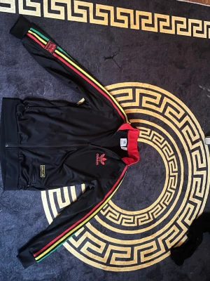 Svart Adidas track jacket XS - Säljer en svart Adidas track jacket med röd krage och färgglada ränder i grönt, gult och rött längs ärmarna. Dragkedja framtill, två fickor och Adidas-logga på bröstet. Perfekt för dig som gillar streetwear och sportig stil.