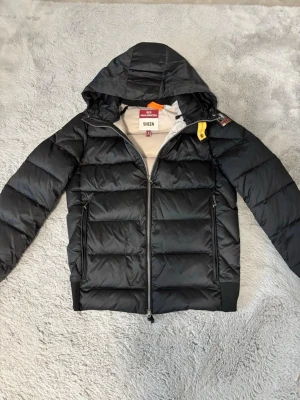 Svart dunjacka från Parajumpers - Snygg svart dunjacka från Parajumpers, modell Sheen. Jackan har huva, två dragkedjeförsedda fickor och klassisk gul detalj vid halsen. Fodrad insida i beige och quiltad design för extra värme. Perfekt för kalla vinterdagar.