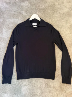 Mörkblå stickad tröja med krage - Snygg mörkblå stickad tröja från These Glory Days i mjuk merinoull. Tröjan har en klassisk krage och långa ärmar, perfekt för en stilren och avslappnad look. Ribbstickade muddar vid ärmslut och nederkant. Tröjan är knappt använd då den inte passar.