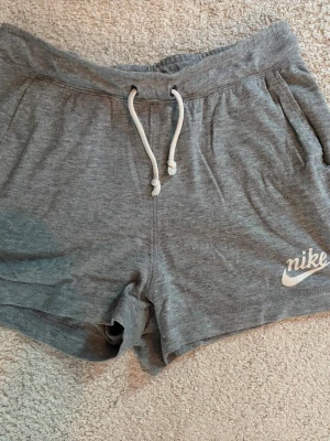 Gråa shorts från Nike med snörning - Gråa mjuka shorts från Nike med vit snörning i midjan och logga på ena benet. Perfekta för chill dagar eller träning. De har en avslappnad passform och är tillverkade i bomullsmix för extra komfort. Skriv pm vid funderingar!☺️