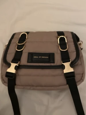 Beige quiltad axelväska Ideal of Sweden - Snygg beige quiltad axelväska från Ideal of Sweden med svarta detaljer och guldfärgade spännen. Väskan har justerbar axelrem och dragkedja upptill. Perfekt storlek för det viktigaste och en stilren look.