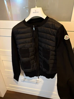 Svart cardiganjacka från Moncler - Snygg svart cardiganjacka från Moncler med quiltad front och stickade ärmar. Jackan har fyra ribbade fickor framtill och dragkedja. Moncler-logga på ena ärmen. Perfekt för lager-på-lager och stilren look. Köpt på vinted med äkthetsverifikationen men knappt använd så i nyskick! Säljes billigare pga lappen i kragen har lossnat