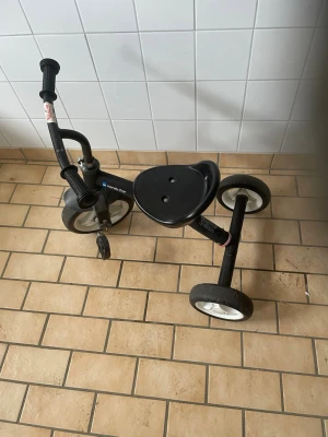 Svart trehjuling för barn - En svart trehjuling med vita hjul och robust ram i metall. Cykeln har ett brett plastformat säte och greppvänliga handtag på styret. Perfekt för yngre barn som vill lära sig cykla och ha kul utomhus.