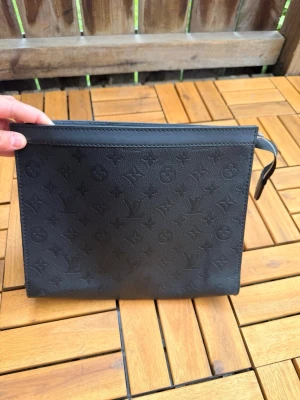 Ny herr Svart kuvertväska med monogram - Stilren svart kuvertväska i läderimitation med präglat monogrammönster över hela ytan. Väskan har dragkedja upptill och en praktisk handledsrem. Insidan har ett större fack och en mindre ficka för småsaker. Perfekt för att bära det viktigaste.