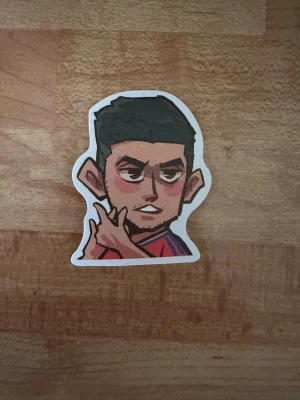 Sticker med tecknad kille - Säljer en sticker med en tecknad kille som har mörkt hår och röd tröja. Han gör ett handtecken och har en självsäker min. Perfekt att sätta på laptop, mobil eller anteckningsbok.