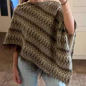 Missoni-inspirerad sicksack-mönstrad poncho topp med fjäderkant / fransar - Superfin och mysig poncho i ett klassiskt Missoni-inspirerat sicksack-mönster i mörkgrå, beige och svarta toner. Unik detalj med fluffiga fjädrar/pälsliknande fransar längs kanten som ger den en riktigt snygg boho-look. Perfekt att slänga över axlarna under kyliga höstdagar eller kvällar!  Fattas lite fransar på ena sidan men inget som märks när den är på.   Storlek ONESIZE, passar i princip alla från XS-XL (har vanligtvis S)