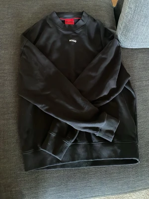 Svart sweatshirt från Hugo - Säljer en svart sweatshirt från Hugo med diskret logga broderad på bröstet. Tröjan är oanvänd. Tröjan har rund halsringning, ribbade muddar vid ärmslut och nederkant samt en clean och stilren look. Perfekt för dig som gillar minimalistisk stil.