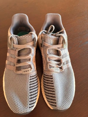 Grå Adidas EQT sneakers - Snygga Adidas EQT sneakers i grått med stickad ovandel och vita detaljer. Skorna har en unik design med ränder på sidan, robust snörning och svart/vit sula med boost-dämpning. Perfekta för dig som gillar streetwear och sportig stil.