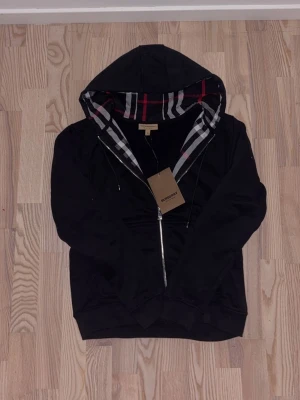 Svart hoodie/Zipper från Burberry med huva - Säljer en svart hoodie från Burberry med dragkedja och klassiskt rutigt foder i huvan. Hoodien har fickor framtill och ribbade muddar. Perfekt för dig som gillar stilrena plagg med exklusiv känsla och snygga detaljer.