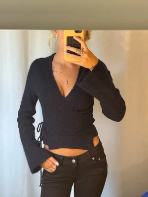 Mörkblå omlottopp med snörning - Säljer en mörkblå omlottopp med v-ringning och långa ärmar. Toppen har snörning i sidan och är croppad i modellen. Materialet ser mjukt och lite fluffigt ut, perfekt för dig som gillar trendiga detaljer.