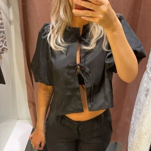 Svart kortärmad topp med knyt - Säljer en snygg svart kortärmad topp med knyt framtill. Den är croppad och har en stilren design som passar perfekt till fest eller en utekväll. Toppen är i ett mjukt material och har en normal passform.
