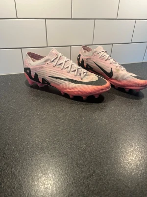 Nike rosa fotbollsskor med dobbar - Säljer ett par Nike fotbollsskor i rosa och svart med stora svarta swoosh-loggor på sidorna. Skorna har snörning, låg profil och dobbar för spel på konstgräs. Ovandelen är i ett lätt syntetmaterial som ger bra bollkontroll och passform.