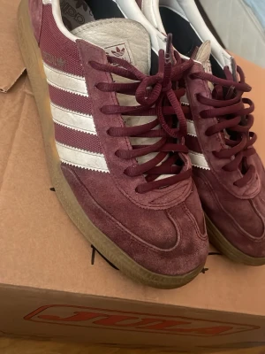 Adidas Handball Spezial vinröda sneakers - Säljer ett par Adidas Handball Spezial sneakers i vinrött mocka med klassiska vita ränder på sidorna och gummisula. Skorna har lågt skaft och retroinspirerad design med Adidas-logga på plösen och guldfärgad text på sidan. Storlek 44… säljer för flyttat precis o behöver plats!