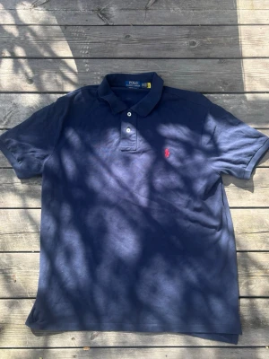 Marinblå piké från Polo Ralph Lauren Oanvänd - Säljer en marinblå pikétröja från Polo Ralph Lauren i custom slim fit. Tröjan har klassisk krage, två knappar och den ikoniska röda logotypen broderad på bröstet. Tillverkad i mjuk bomull, perfekt för en clean och stilren look. Oanvänd