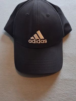 Svart keps från Adidas - Snygg svart keps från Adidas med klassisk böjd skärm och justerbart spänne bak. Framtill sitter den ikoniska Adidas-loggan broderad i vitt. Tillverkad i ett lätt och bekvämt material som passar perfekt till streetwear och sportiga outfits. Storlek L Fit the most. Färgen kan vara lite annorlunda än bilden.