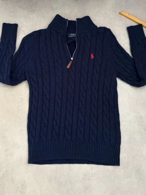 Marinblå kabelstickad tröja Polo - Kabelstickad marinblå tröja från Polo Ralph Lauren med halv dragkedja och hög krage. Tröjan har ett rött broderat logomärke på bröstet och ribbade muddar vid ärmslut och nederkant. Perfekt för kyliga dagar.Priset kan diskuteras 🍾