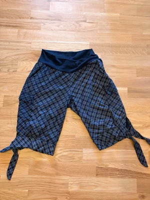 Svarta rutiga långa shorts med knytband - Snygga svarta shorts med rutmönster och bred, mjuk midja. Shortsen har knytband nertill på benen och praktiska fickor på sidorna. Väldigt nopprigt material. Dessutom står ingen strl så jag gissar på M