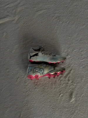 Puma fotbollsskor med rosa dobbar - Snygga fotbollsskor från Puma i vit med svarta och ljusblåa detaljer och rosa dobbar. Skorna har snörning, låg profil och en stor svart Puma-logga på sidan samt TR-tryck på hälen. Perfekta för dig som vill sticka ut på planen.