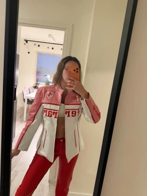 Vit och rosa Ferrari skinnjacka - Säljer en unik vit och rosa skinnjacka från Ferrari med röda detaljer och broderade loggor. Jackan har dragkedja framtill, ståkrage och texten 'Ferrari' i rött över bröstet och på ärmen. Snygg passform och cool racingstil.